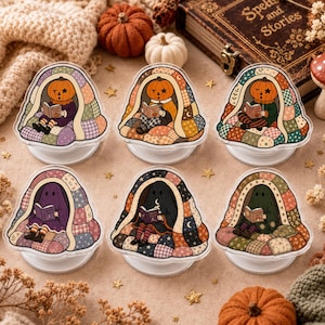 May include: Six circular phone grips featuring illustrations of pumpkin-headed characters and ghosts reading books while sitting under colorful patchwork quilts. Each design showcases unique patterns and autumn-inspired color palettes.
