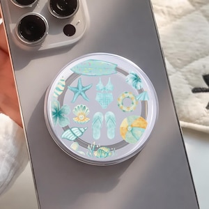 May include: A clear phone grip with a beach-themed design, featuring a surfboard, starfish, swimsuit, and other summer elements in shades of blue, green, and yellow. The grip is attached to a gray smartphone.