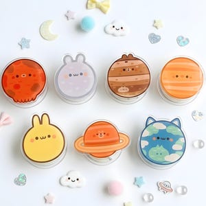 May include: A collection of clear, round, decorative containers with cartoon planet and animal designs. The containers are orange, brown, yellow, blue, and gray. Decorative stars, clouds, and bows surround the containers.