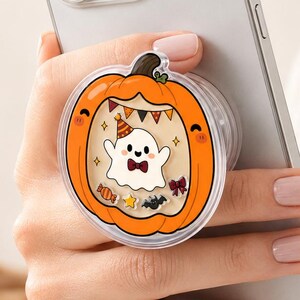 May include: A silver smartphone featuring a circular phone grip on the back. The grip is shaped like an orange pumpkin with a clear window containing a small white ghost wearing a party hat and bow tie, surrounded by tiny festive Halloween icons.