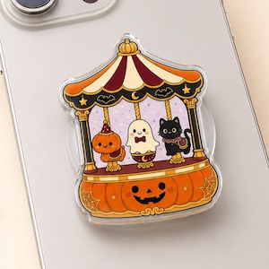 May include: A silver smartphone with a circular phone grip attached to the back. The grip features a colorful illustration of a carousel with a pumpkin, a ghost, and a black cat, all set upon a base shaped like a large orange jack-o'-lantern.