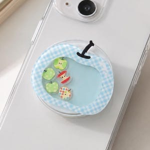 May include: A clear phone case with a decorative apple-shaped phone grip. The grip features a light blue background with a gingham border and small apple designs in green, red, and yellow.