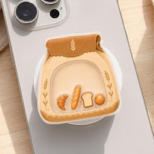 May include: A light gray smartphone with a circular phone grip attached to the back. The grip features a tan paper bag illustration with a wheat stalk icon and small graphics of a croissant, a baguette, a slice of bread, and a round bun.