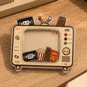 May include: A clear acrylic shaker charm shaped like a vintage television set. Inside the frame are small floating charms of a VHS tape, popcorn, a remote control, and a bag of chips. The television features printed details like REC, VHS, and a smiley face.