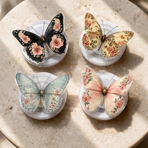 May include: Four butterfly-shaped phone grips with floral designs. The butterflies have black, yellow, blue, and pink wings, each with a unique floral pattern. They are mounted on white circular bases, arranged on a light-colored, round surface.