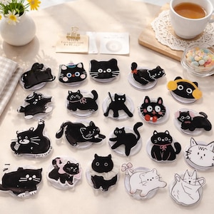May include: A collection of cat-themed phone grips in various poses and designs. The grips are primarily black and white, with some featuring bows, hats, and other decorative elements. The collection is arranged on a light-colored surface.