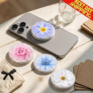 May include: A collection of floral phone grips featuring a variety of flower designs, including a pink peony, blue daisy, white cosmos, and a light purple flower with a yellow center. The grips are on a silver smartphone.