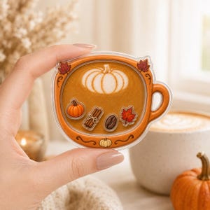 May include: A clear acrylic pin shaped like a coffee mug filled with brown liquid. The mug features a white pumpkin latte art design, a small orange pumpkin, a cinnamon stick bundle, a dark coffee bean, and a red maple leaf graphic.