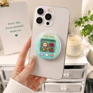 May include: A light-colored smartphone with a teal pop socket featuring a pixelated design of a small character, heart, star, and cherries. The pop socket has the text "TAMA BABE". The phone is held in a person's hand.