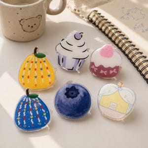 May include: A collection of colorful, clear acrylic pins in various fruit and dessert shapes. Designs include a yellow pear, a blue blueberry, a cupcake, a strawberry-topped dessert, and a bear-themed apple.
