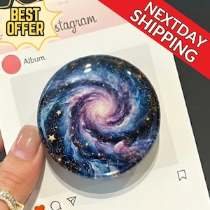 May include: A round phone grip with a galaxy design featuring swirling blues, purples, and gold stars. The grip is held over a white surface with the Instagram logo visible. The words "BEST OFFER" and "NEXTDAY SHIPPING" are also visible.