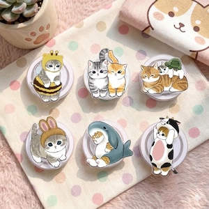 May include: A collection of cat-themed phone grips. Each grip features a cartoon cat in various costumes, including a bee, bunny, shark, and cow. The grips are round and have a white base with a clear top.