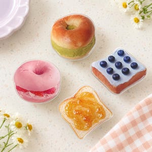 May include: Four food-shaped coasters: a bagel with green filling, a pink donut with red fruit, a square toast slice with blue icing and nine blueberries, and a toast slice with orange marmalade. Each item is encased in a clear plastic shell.