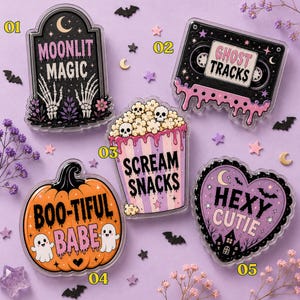 May include: Five acrylic pins featuring spooky designs: a tombstone labeled MOONLIT MAGIC, a cassette tape labeled GHOST TRACKS, a popcorn bucket labeled SCREAM SNACKS, an orange pumpkin labeled BOO-TIFUL BABE, and a heart labeled HEXY CUTIE.