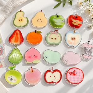 May include: A collection of colorful fruit-shaped acrylic charms. The charms include pears, apples, strawberries, and peaches, each with unique designs and details. Some have smiling faces or cute expressions. The charms are clear with vibrant colors.