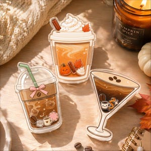 May include: Three acrylic charms featuring beverage illustrations: a pumpkin spice latte with whipped cream and cinnamon, an iced coffee in a plastic cup with a pink bow and green straw, and an espresso martini with coffee beans and a small mug.