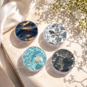 May include: Four round, clear containers with decorative designs. One features a water scene with golden light, another has butterfly patterns, a third shows a blue mosaic with a flower, and the last depicts rippling water. The containers are on a light-colored surface.