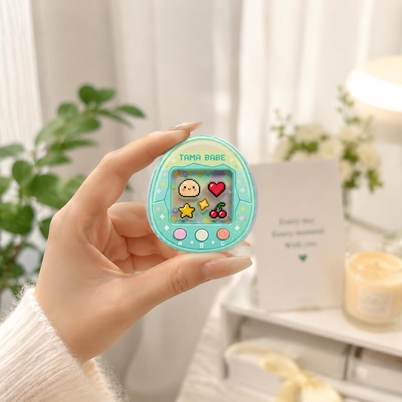May include: A mint green handheld digital pet game with the text "TAMA BABE" at the top. The screen displays pixelated icons of a smiling face, heart, star, and cherries. The game has three colored buttons at the bottom.