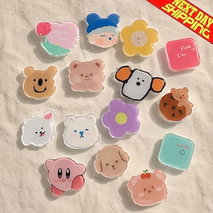 May include: A collection of colorful, cartoon-style acrylic charms in various shapes, including bears, flowers, and characters. Some charms have text like "Fine I'm" and "Luck". The charms are on a cream-colored surface.