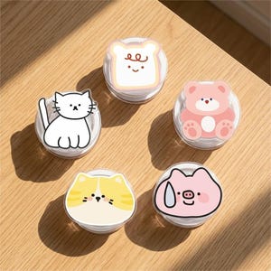 May include: A collection of five cartoon-style phone grips. Designs include a white cat, a slice of toast, a pink bear, a yellow and white cat, and a pink pig. Each grip is round and has a clear plastic base.