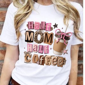 May include: White t-shirt with the text "Half Mom Half Coffee" in a mix of fonts and patterns. The design includes a coffee cup with a pink bow and star accents. The shirt is a casual wear item.