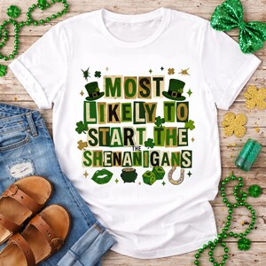 May include: White t-shirt with the phrase "MOST LIKELY TO START THE SHENANIGANS" in green and brown lettering. The design includes shamrocks, a leprechaun hat, and other St. Patrick's Day themed graphics. The shirt is paired with blue jeans and sandals.