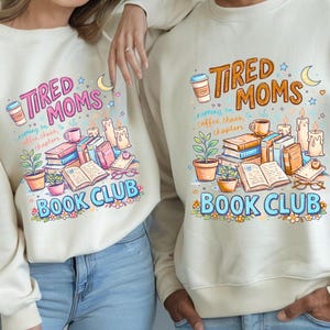 Puede incluir: Sudadera de color crema con un gráfico colorido. El gráfico incluye el texto "TIRED MOMS BOOK CLUB" con ilustraciones de libros, tazas de café, velas y plantas. La sudadera es de estilo casual.