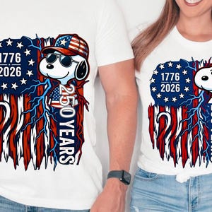 May include: White t-shirts featuring a patriotic design with Snoopy wearing sunglasses and a hat. The design includes the American flag, the years 1776 and 2026, and the text "250 YEARS".