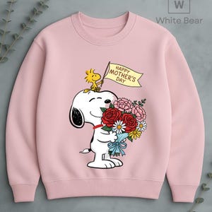 May include: A light pink sweatshirt featuring a Snoopy graphic holding a bouquet of flowers and a banner that reads "Happy Mother's Day." A yellow bird is perched on Snoopy's head. The sweatshirt has a crew neck and long sleeves.