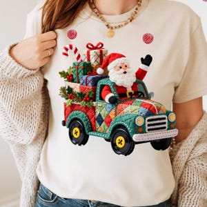 May include: A cream-coloured t-shirt with a festive Christmas design. The graphic shows Santa Claus driving a patchwork truck loaded with presents, holly, and a candy cane. Red and green colours create a cheerful holiday theme.