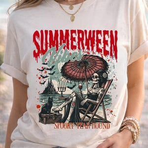 May include: Cream-colored t-shirt with a graphic featuring a skeleton relaxing on a beach chair under a red umbrella, holding a glass of wine. The text reads "SUMMERWEEN Vibes" and "SPOOKY YEAR ROUND". A black cat sits nearby.