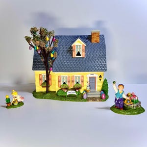 May include: A miniature Easter village scene featuring a yellow house with a blue roof, a tree with colorful Easter egg ornaments, and figurines of people and bunnies. The house has a sign that says "Happy Easter".