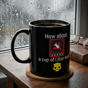 May include: A black ceramic mug featuring the white text How about a cup of Liber-tea? with a red square graphic containing a white bomb icon and directional arrows, plus a yellow skull icon printed on the front surface of the cylindrical vessel.