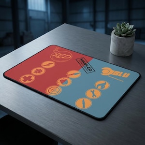 Op de afbeelding: Een rechthoekige muismat met een rood en blauw gesplitst ontwerp. De rode kant heeft het woord "RED" en diverse oranje pictogrammen. De blauwe kant heeft het woord "BLU" en vergelijkbare pictogrammen. De muismat heeft ook de tekst "MANN CO."