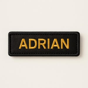 Op de afbeelding: Zwart rechthoekig naamlabel met de naam "ADRIAN" geborduurd in goudkleurig garen. Het label heeft een gestikte rand en is geplaatst tegen een neutrale achtergrond.