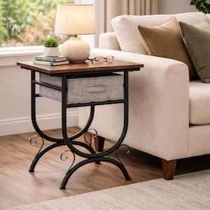 May include: A small, dark wood side table with a black metal frame and a gray fabric drawer. A cream-colored lamp with a beige lampshade sits on top, along with a small plant, books, and eyeglasses. The table is next to a beige sofa.