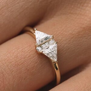 May include: A gold ring with two large, triangular white gemstones and smaller square-cut stones. The ring has a thin, polished gold band. The ring is displayed on a person's finger.