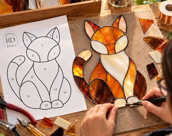 May include: A stained glass fox suncatcher in progress, featuring amber, orange, and cream glass pieces joined by dark solder lines. Beside it lies a numbered paper pattern labeled RD Designs, with glass cutting tools and loose glass shards nearby.
