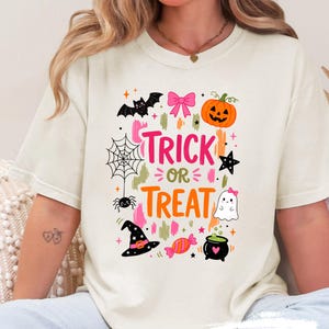 May include: An ivory t-shirt featuring a colourful Halloween graphic with the text "TRICK OR TREAT". The design includes a black bat, a pink bow, an orange pumpkin, a spider web, a spider, a black star, a ghost, a witch hat, candy, and a cauldron.