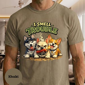 Puede incluir: Camiseta caqui con un gráfico de tres perros de dibujos animados: un husky con gafas de sol, un bulldog con un palo de dinamita y un corgi. El texto "I SMELL TROUBLE" está encima de los perros.
