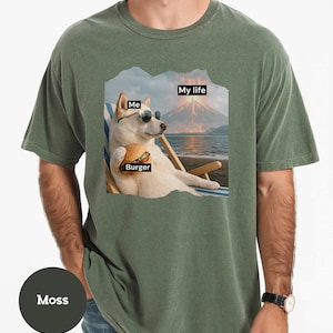 May include: Moss green t-shirt featuring a graphic of a dog wearing sunglasses, holding a burger, and relaxing on a beach chair. The graphic includes the text "Me", "Burger", and "My life".