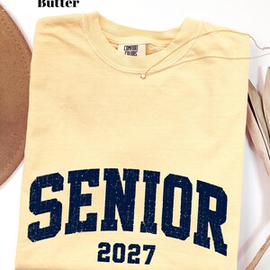 Pode incluir: Uma t-shirt de manga curta na cor amarelo manteiga com gola redonda e etiqueta da marca Comfort Colors. A frente apresenta o texto SENIOR em azul marinho escuro com uma fonte universit&aacute;ria desgastada e o n&uacute;mero 2027 impresso abaixo.