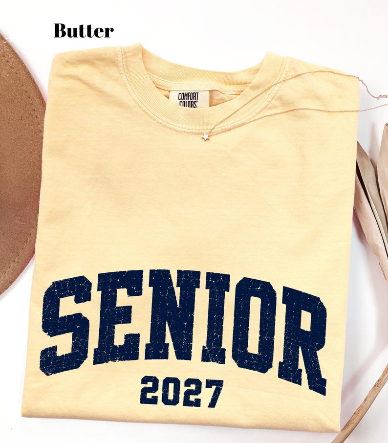 Pode incluir: Uma t-shirt de manga curta na cor amarelo manteiga com gola redonda e etiqueta da marca Comfort Colors. A frente apresenta o texto SENIOR em azul marinho escuro com uma fonte universit&aacute;ria desgastada e o n&uacute;mero 2027 impresso abaixo.