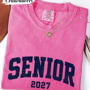 Pode incluir: Uma t-shirt de manga curta cor-de-rosa brilhante com um gr&aacute;fico azul-marinho desgastado que diz SENIOR 2027. A pe&ccedil;a apresenta uma etiqueta da marca Comfort Colors no decote interior e um pequeno colar com pendente circular dourado.