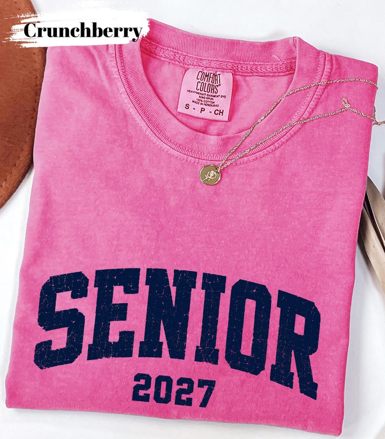 Pode incluir: Uma t-shirt de manga curta cor-de-rosa brilhante com um gr&aacute;fico azul-marinho desgastado que diz SENIOR 2027. A pe&ccedil;a apresenta uma etiqueta da marca Comfort Colors no decote interior e um pequeno colar com pendente circular dourado.