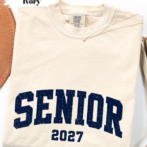 Pode incluir: Uma t-shirt de manga curta na cor marfim com gola redonda e uma etiqueta da marca Comfort Colors no interior da gola. A frente apresenta a palavra SENIOR numa fonte universit&aacute;ria azul-marinho desgastada, com o n&uacute;mero 2027 impresso abaixo.