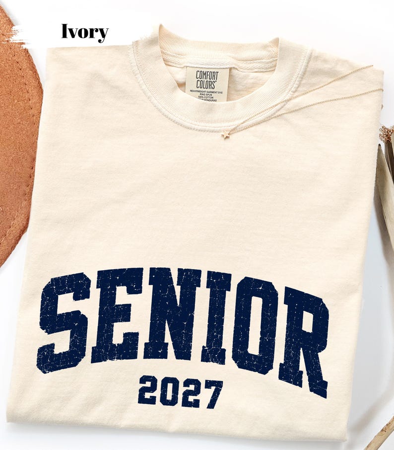 Pode incluir: Uma t-shirt de manga curta na cor marfim com gola redonda e uma etiqueta da marca Comfort Colors no interior da gola. A frente apresenta a palavra SENIOR numa fonte universit&aacute;ria azul-marinho desgastada, com o n&uacute;mero 2027 impresso abaixo.