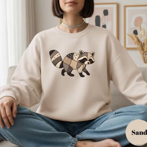 May include: A sand-colored crewneck sweatshirt featuring a central graphic of a raccoon designed with a patchwork pattern. The raccoon illustration incorporates various shades of brown, beige, and cream in a quilted style with visible stitching.