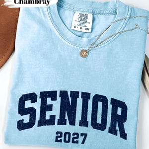 Pode incluir: Uma t-shirt de manga curta azul-clara com um gr&aacute;fico azul-marinho desgastado que diz SENIOR 2027. A pe&ccedil;a apresenta uma etiqueta da marca Comfort Colors no colarinho indicando o tamanho S e um colar de corrente prateada com um pequeno pendente.