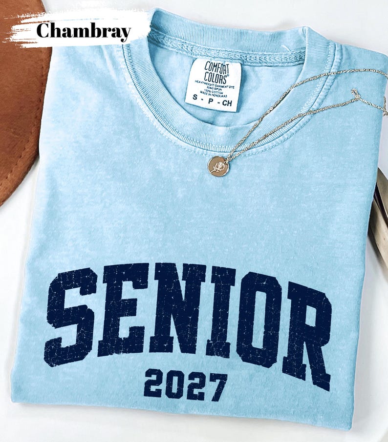 Pode incluir: Uma t-shirt de manga curta azul-clara com um gr&aacute;fico azul-marinho desgastado que diz SENIOR 2027. A pe&ccedil;a apresenta uma etiqueta da marca Comfort Colors no colarinho indicando o tamanho S e um colar de corrente prateada com um pequeno pendente.